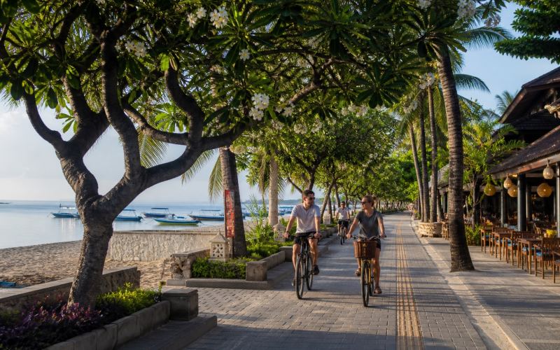Cyclists on a shady promenade with cafes and sea views in Sanur